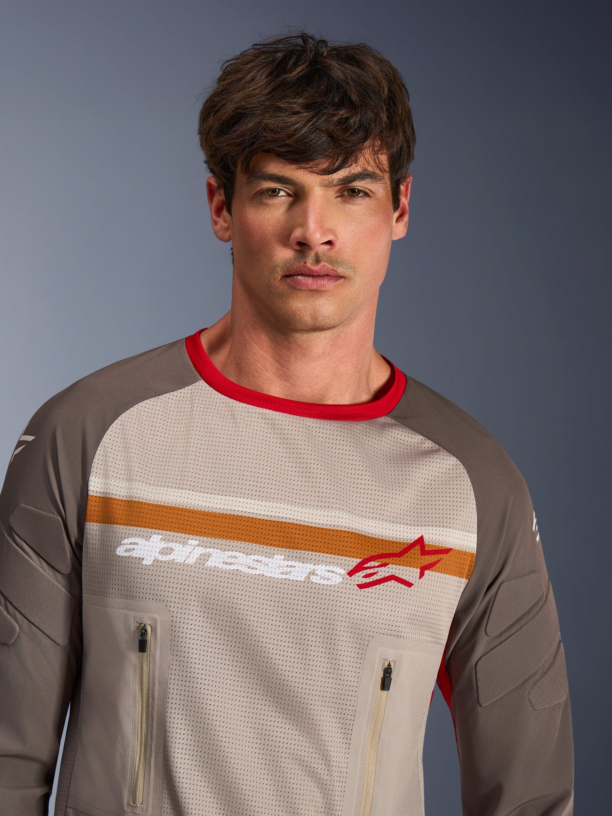 Male model wearing Alpinestars Maxdura Dual Jersey in Aluminum Walnut, adventure motorcycle jersey with full mesh torso, abrasion-resistant sleeves, red collar, and zippered chest pockets, featuring orange and white chest stripes with Alpinestars logo.