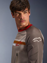 Male model wearing Alpinestars Maxdura Dual Jersey in Aluminum Walnut, featuring a ventilated mesh torso, red collar, and orange chest stripe. Close-up view of abrasion-resistant sleeve with integrated padding and a zippered chest pocket for adventure riding.