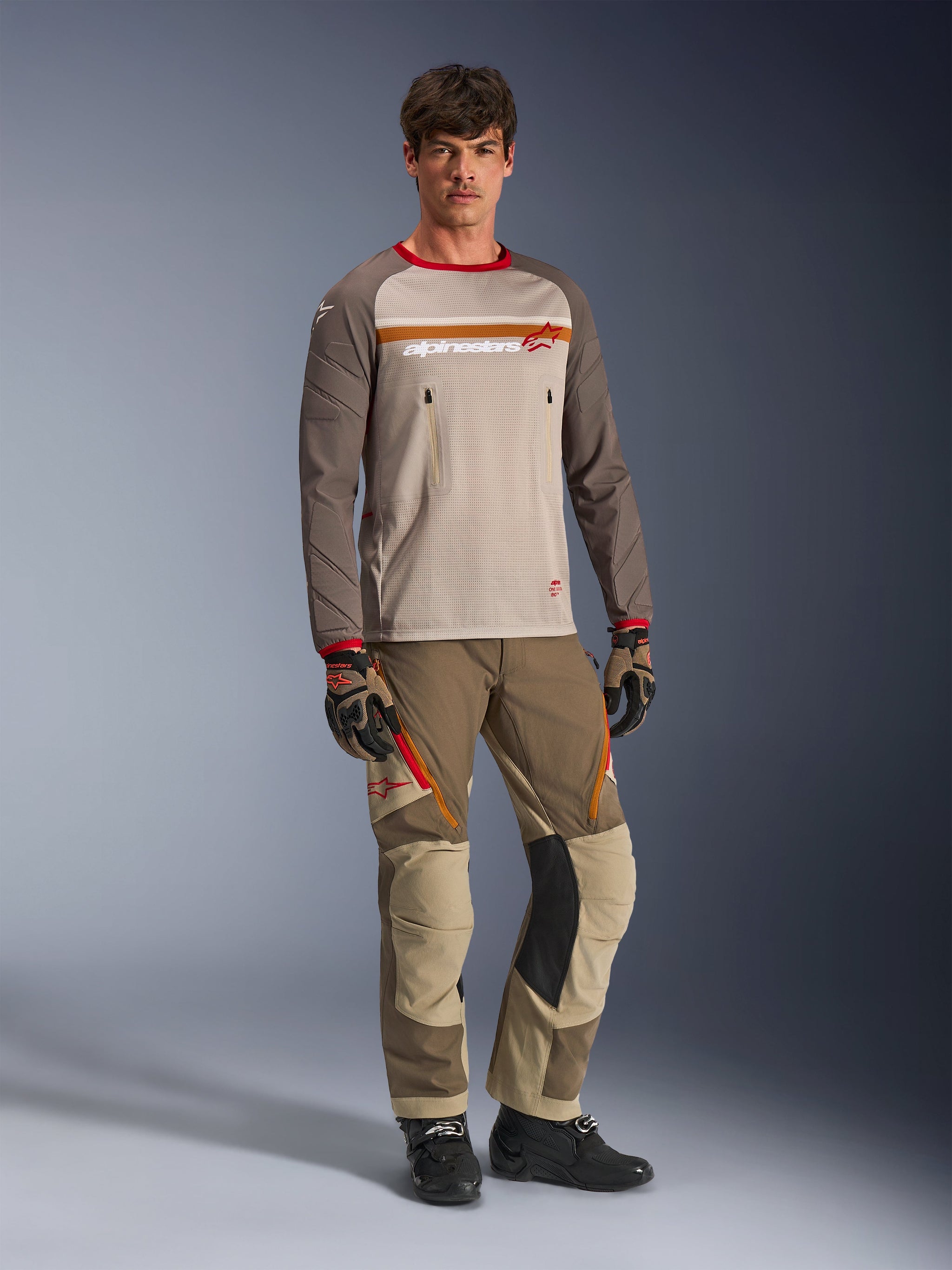 Model wearing the Alpinestars Maxdura Dual Jersey in Aluminum Walnut, featuring a grey mesh torso, brown sleeves with red cuffs, and zippered chest pockets, styled with matching off-road gear for adventure riding.