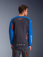 Alpinestars Maxdura Dual Jersey in Deep Gray and Royal Blue, back view of male model, mesh torso construction with orange Astars logo, long sleeves with white branding, maroon collar and cuffs, worn with gray technical pants, dual-sport and adventure motorcycle gear