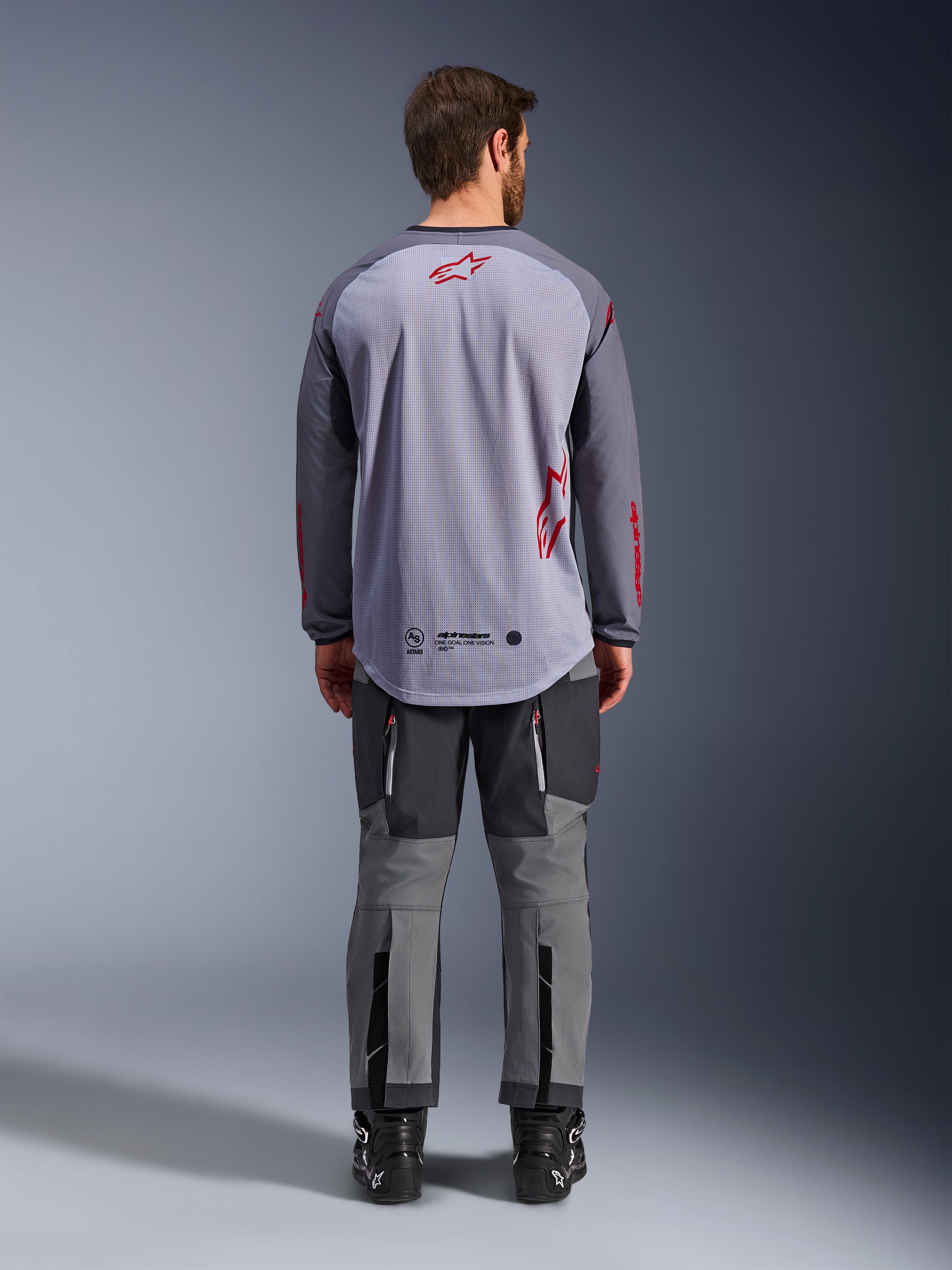 Male model from behind wearing the Alpinestars Maxdura Dual Jersey, Mid Gray and Dark Gray adventure jersey with red logo accents, mesh back construction, worn with matching gray off-road pants and black boots