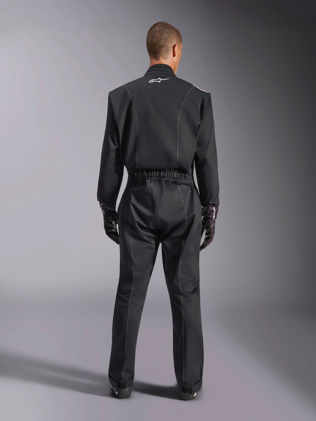 Alpinestars Kart Indoor V2 Suit, Black Karting Suit, rear full-body view on model, featuring white piping details and a small white Astars logo on the back collar, elastic waist belt, worn with black racing gloves, one-piece motorsport apparel.