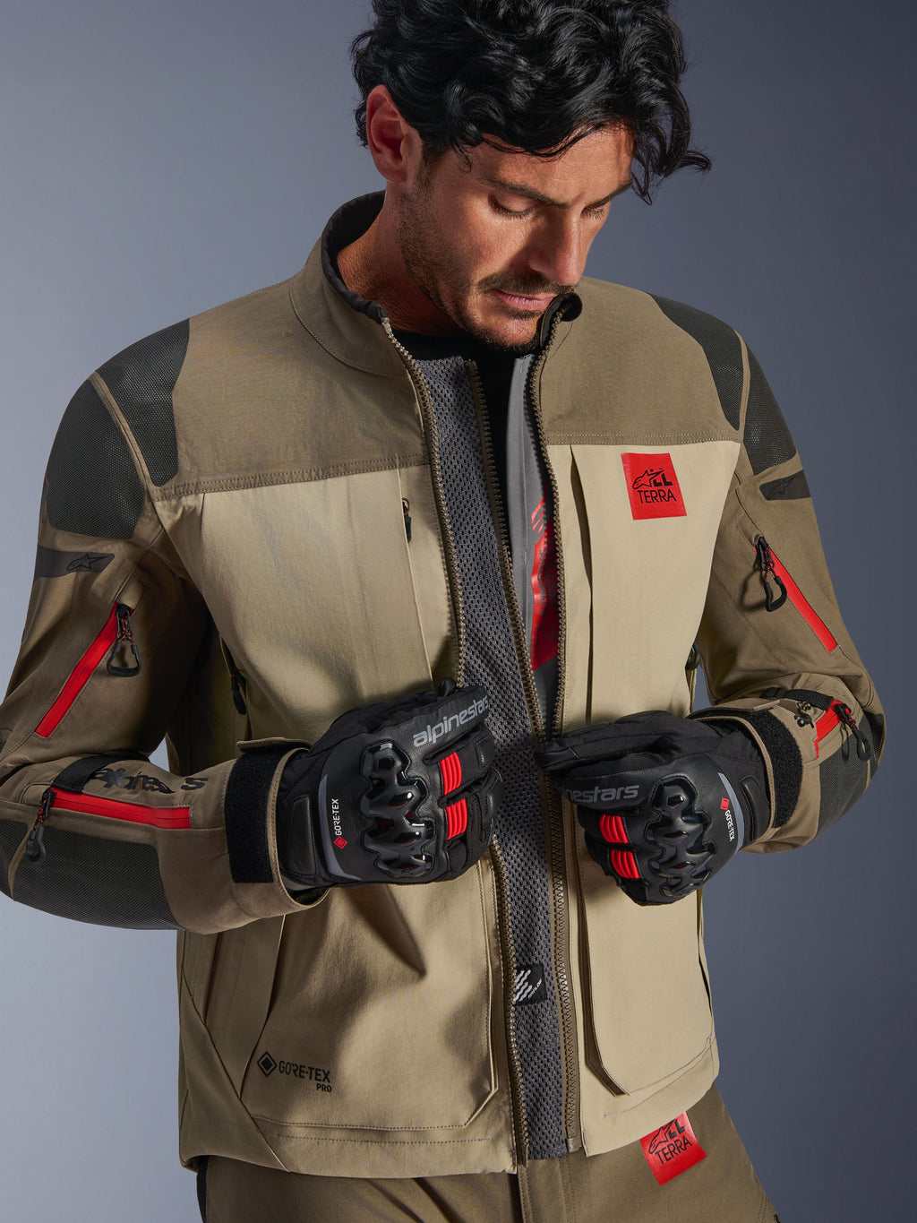 Alpinestars Nazca 3L GORE-TEX PRO Jacket, Walnut Rock Brown, adventure motorcycle jacket with unzipped front revealing central mesh airflow panel, red All Terra branding, worn with black GORE-TEX riding gloves, technical gear for off-road touring