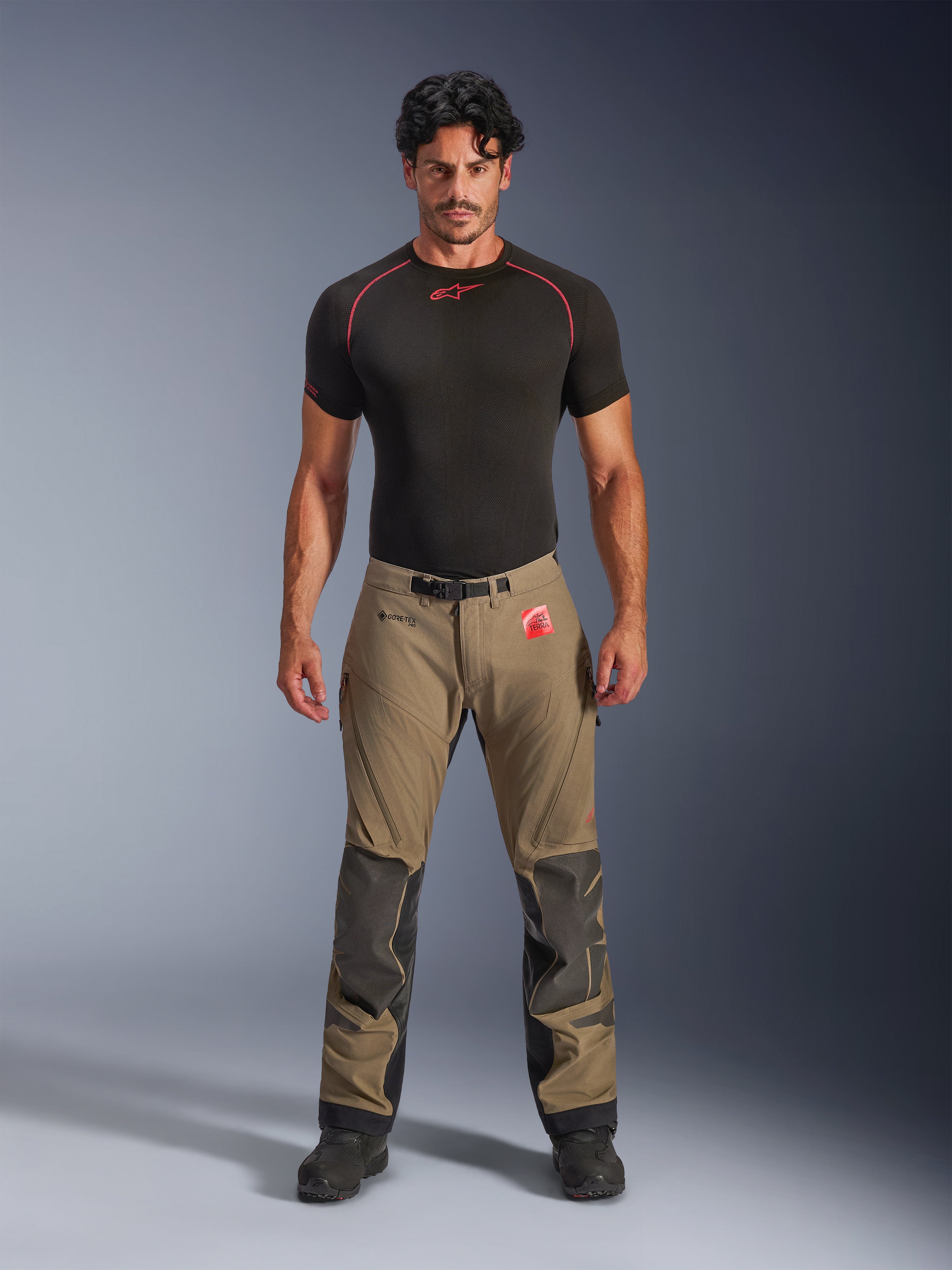 Male model wearing Alpinestars Nazca 3L GORE-TEX PRO Pants in walnut brown and black, technical adventure riding gear featuring Superfabric knee reinforcements and a magnetic waist buckle, paired with a black Alpinestars base layer shirt, studio front view.