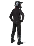 Enduro rider viewed from the back wearing the Alpinestars Lite-Dura Softshell Jacket in Black, paired with black technical riding pants and boots, featuring a zippered rear storage pocket and Alpinestars logo on the lower hem, complete off-road gear for motocross.