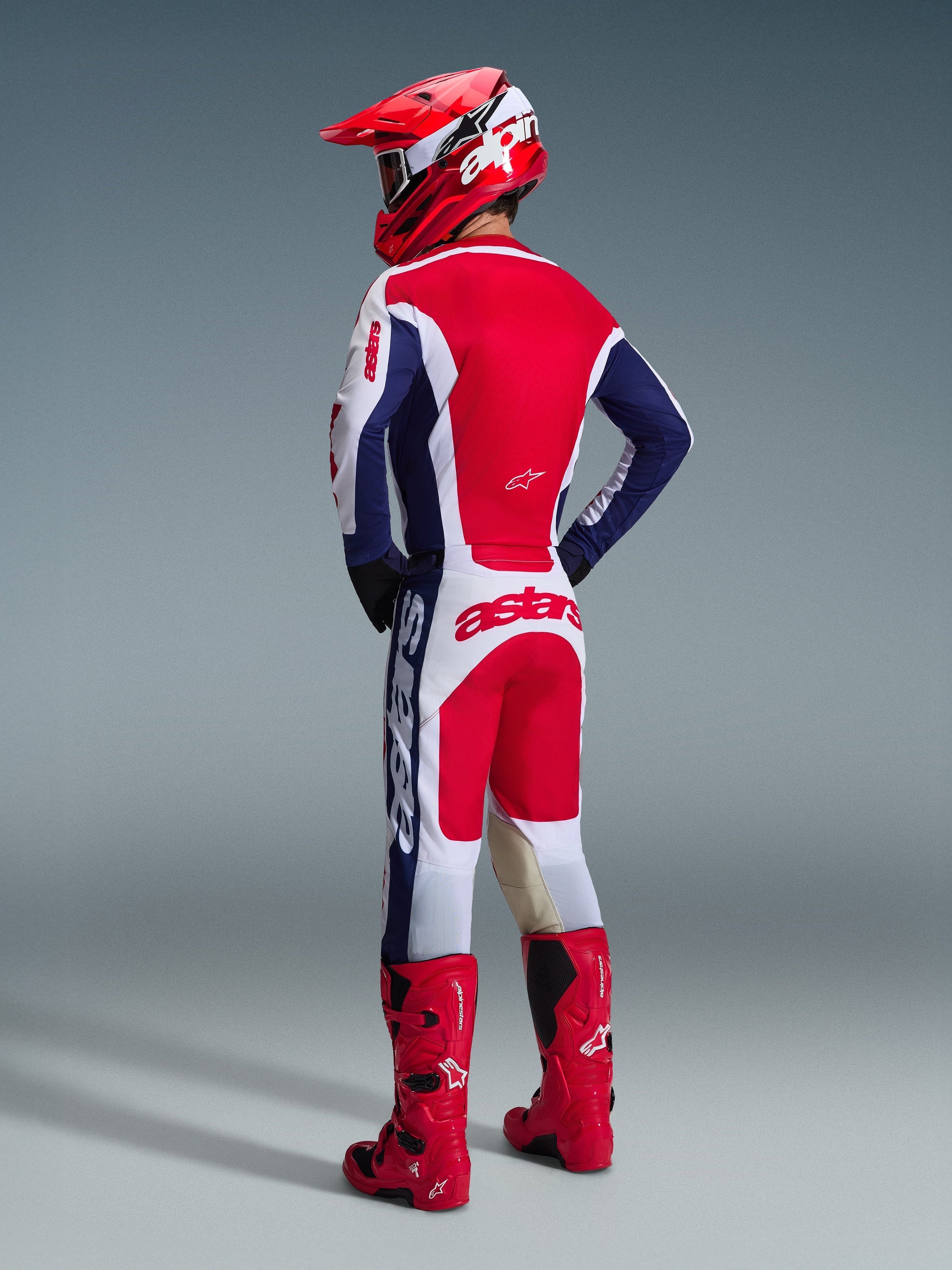 Motocross rider viewed from behind wearing Alpinestars 2026 Racer Riway Pants in White, Blue, and Red, paired with a matching jersey, red helmet, and bright red boots, showcasing the bold branding on the seat and athletic off-road fit.