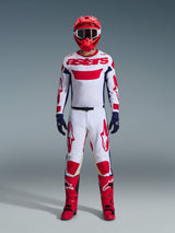 Motocross rider wearing complete racing gear, white jersey with red Alpinestars branding and blue accents, Alpinestars 2026 Racer Riway Pants in White Blue Red, red helmet with matching goggles, red and black boots, navy blue gloves, coordinated outfit for off-road racing