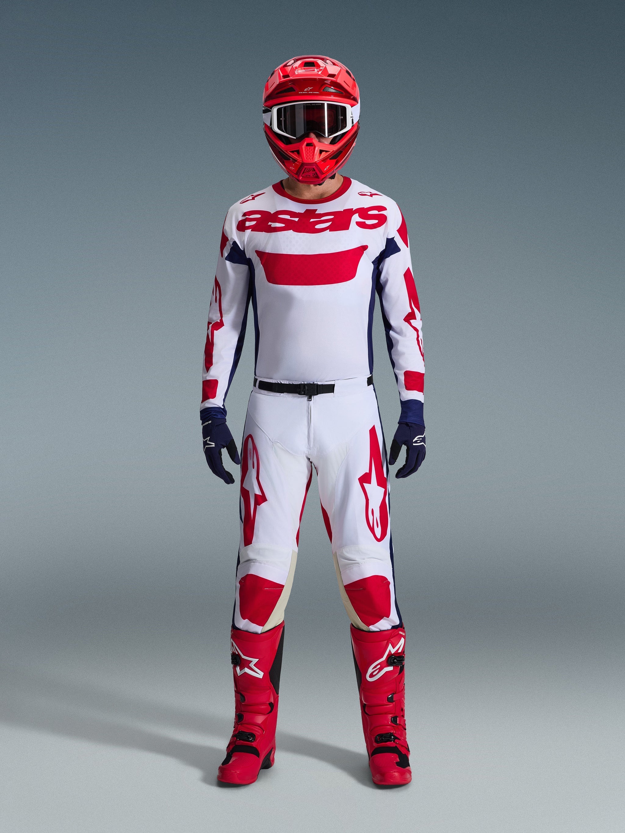 Motocross rider wearing complete racing gear, white jersey with red Alpinestars branding and blue accents, Alpinestars 2026 Racer Riway Pants in White Blue Red, red helmet with matching goggles, red and black boots, navy blue gloves, coordinated outfit for off-road racing