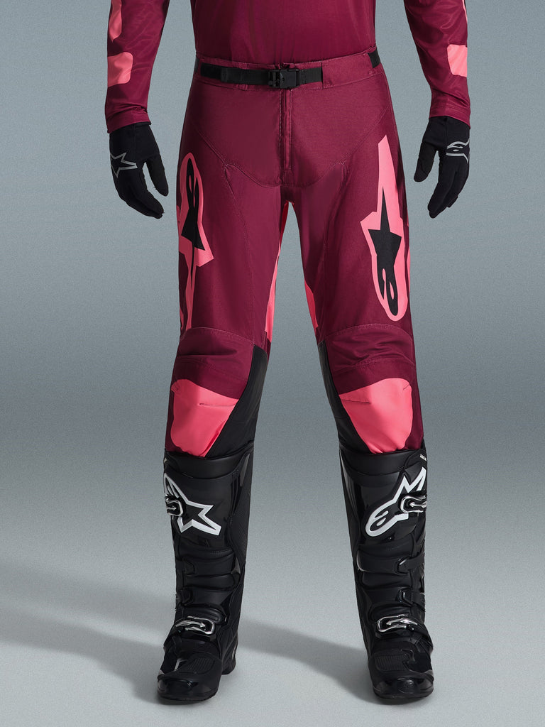 Alpinestars 2026 Racer Riway Pants, Motocross Pants, Dark Red Coral, worn with black gloves and boots, featuring large coral Alpinestars logos on thighs, black waist buckle, and pre-curved leg design for off-road racing