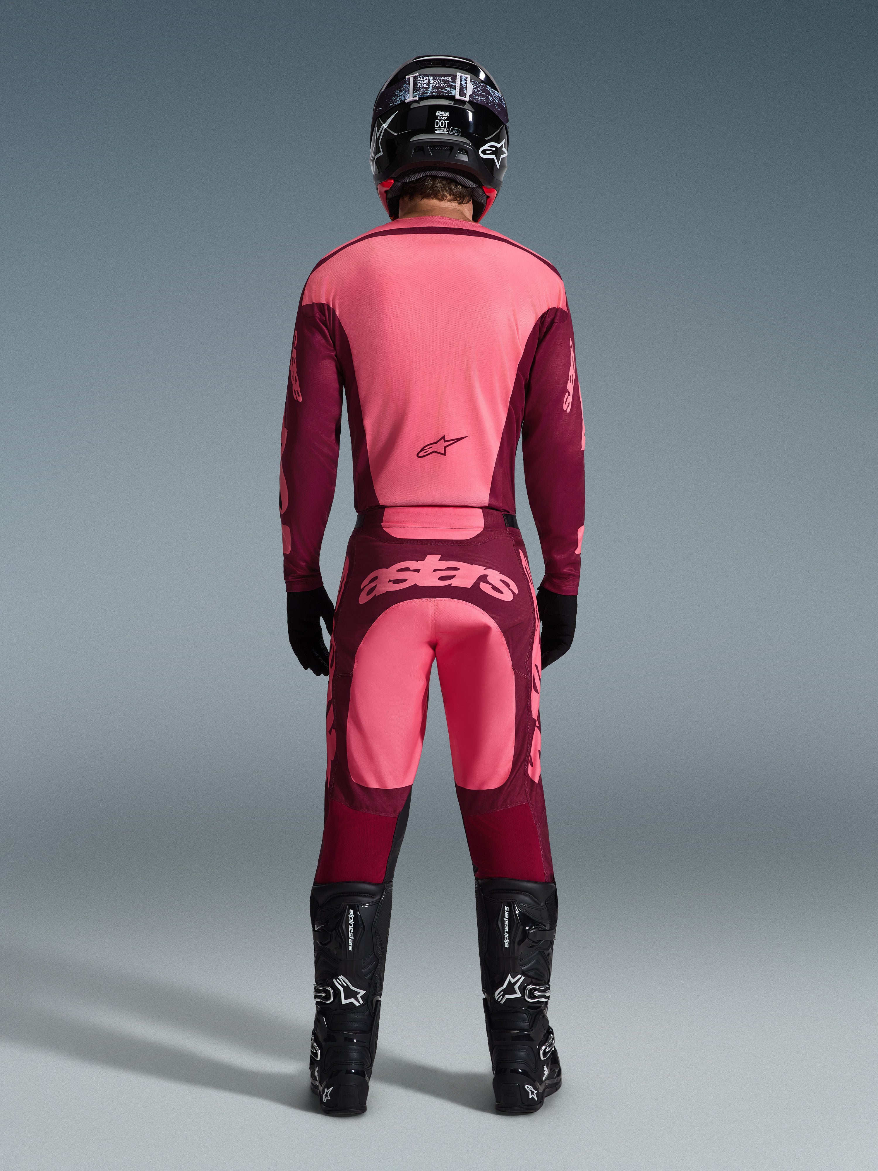 Rear view of a motocross rider wearing the 2026 Racer Riway Gear Set, pink and burgundy jersey, matching burgundy pants with light pink 'astars' logo on the seat, black helmet, black boots, and black gloves, coordinated outfit for off-road performance