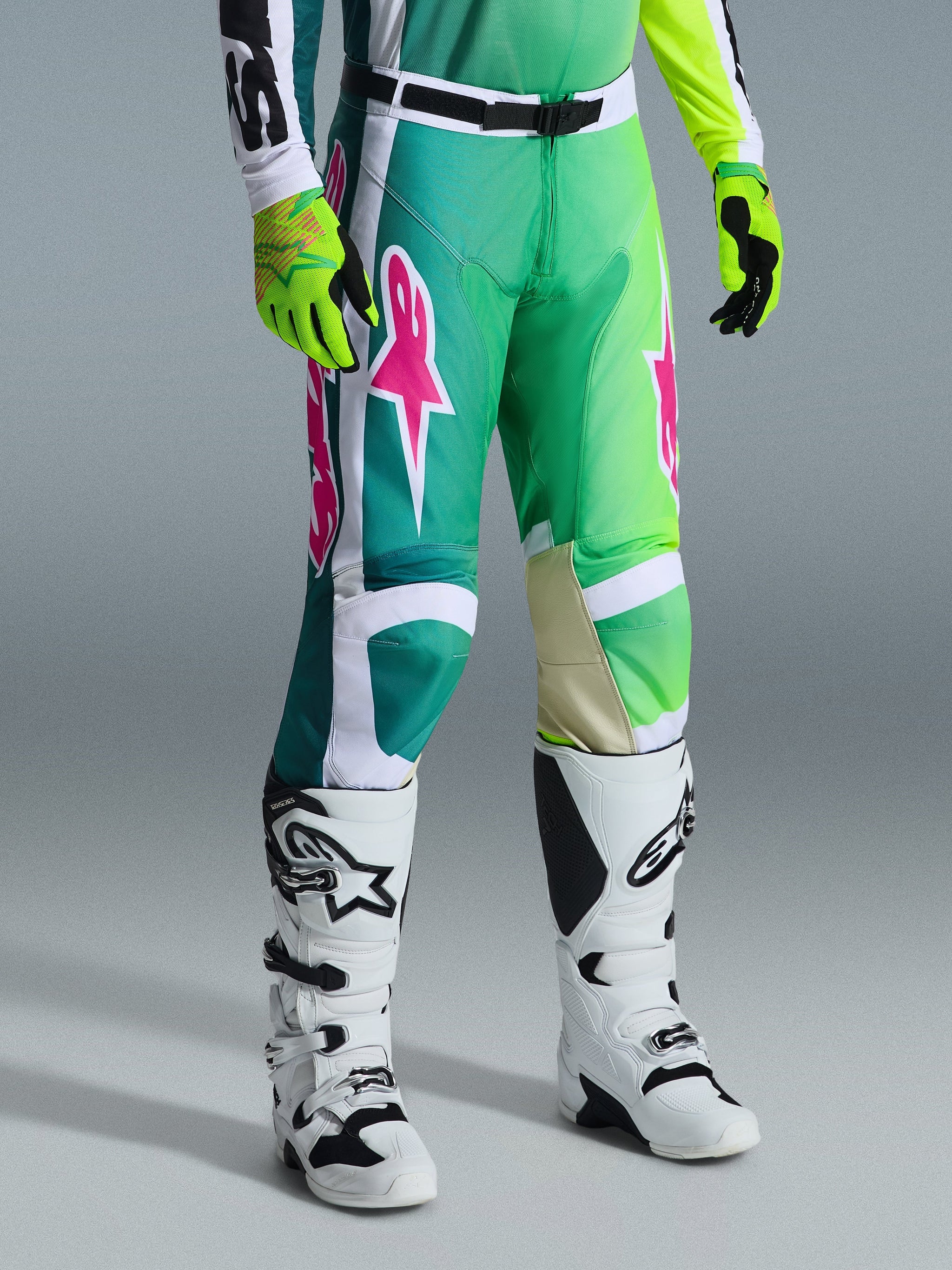 Alpinestars 2026 Racer Portl Pants, Motocross Pants, Green White and Black with pink logos, worn with white boots and neon yellow gloves, featuring bold graphic patterns and an adjustable black waist buckle, performance gear for off-road riding.