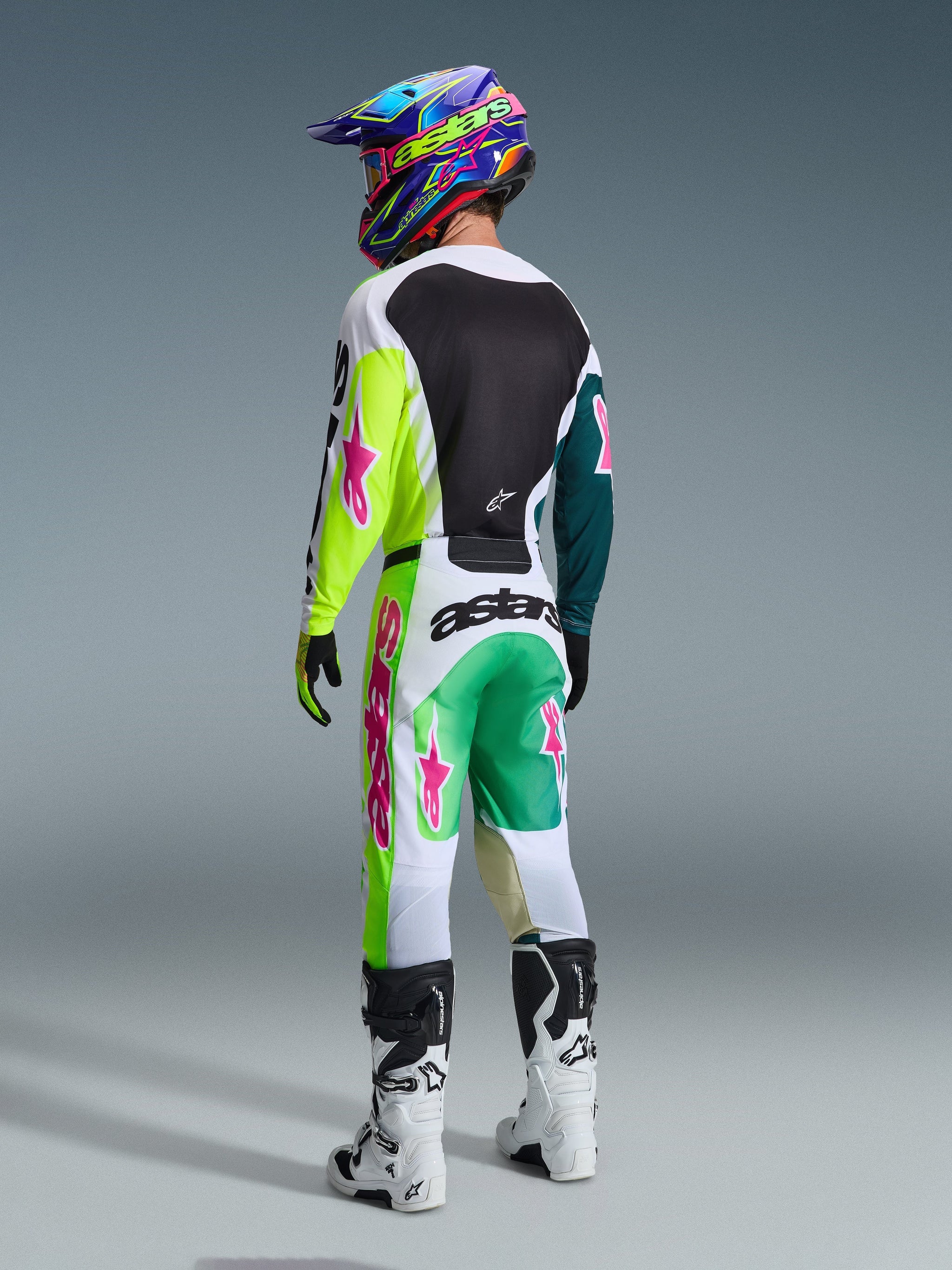 Motocross rider from behind wearing Alpinestars 2026 Racer Portl Pants in Green White Black, matching jersey with lime and pink accents, colorful helmet, white and black boots, full off-road racing gear set.