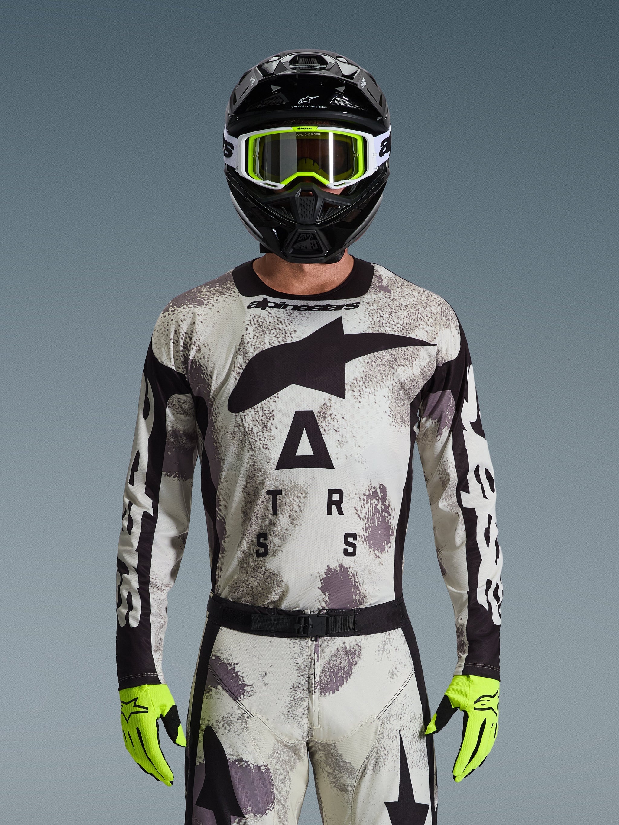 Motocross rider wearing Alpinestars 2026 Racer Lahnd Jersey, Sand Camo pattern in gray and white, large black Astar logo and branding on the chest, paired with a black helmet, neon yellow goggles and gloves, performance gear for off-road riding