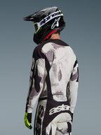 Motocross rider from the back wearing the Alpinestars 2026 Racer Lahnd Gear Set, Gray and White Sand Camo jersey and pants with black accents, black helmet with white Alpinestars logo, neon yellow and black gloves, coordinated off-road performance outfit