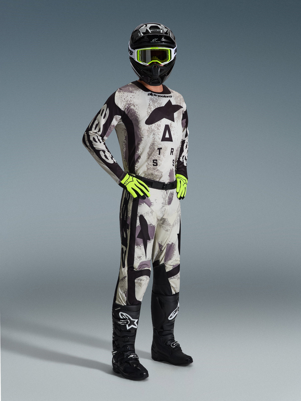 Motocross rider wearing the 2026 Racer Lahnd Jersey in Sand Camo, gray and black camouflage pattern with Alpinestars branding, matching motocross pants, neon yellow gloves, black helmet with yellow goggles, and black boots, complete professional off-road racing outfit.