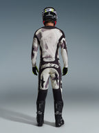 Motocross rider from behind wearing the Alpinestars 2026 Racer Lahnd Pants in Sand Camo, matching long-sleeve jersey with gray camouflage pattern, black motocross boots, black helmet, and "astars" branding on the seat, designed for professional off-road racing.