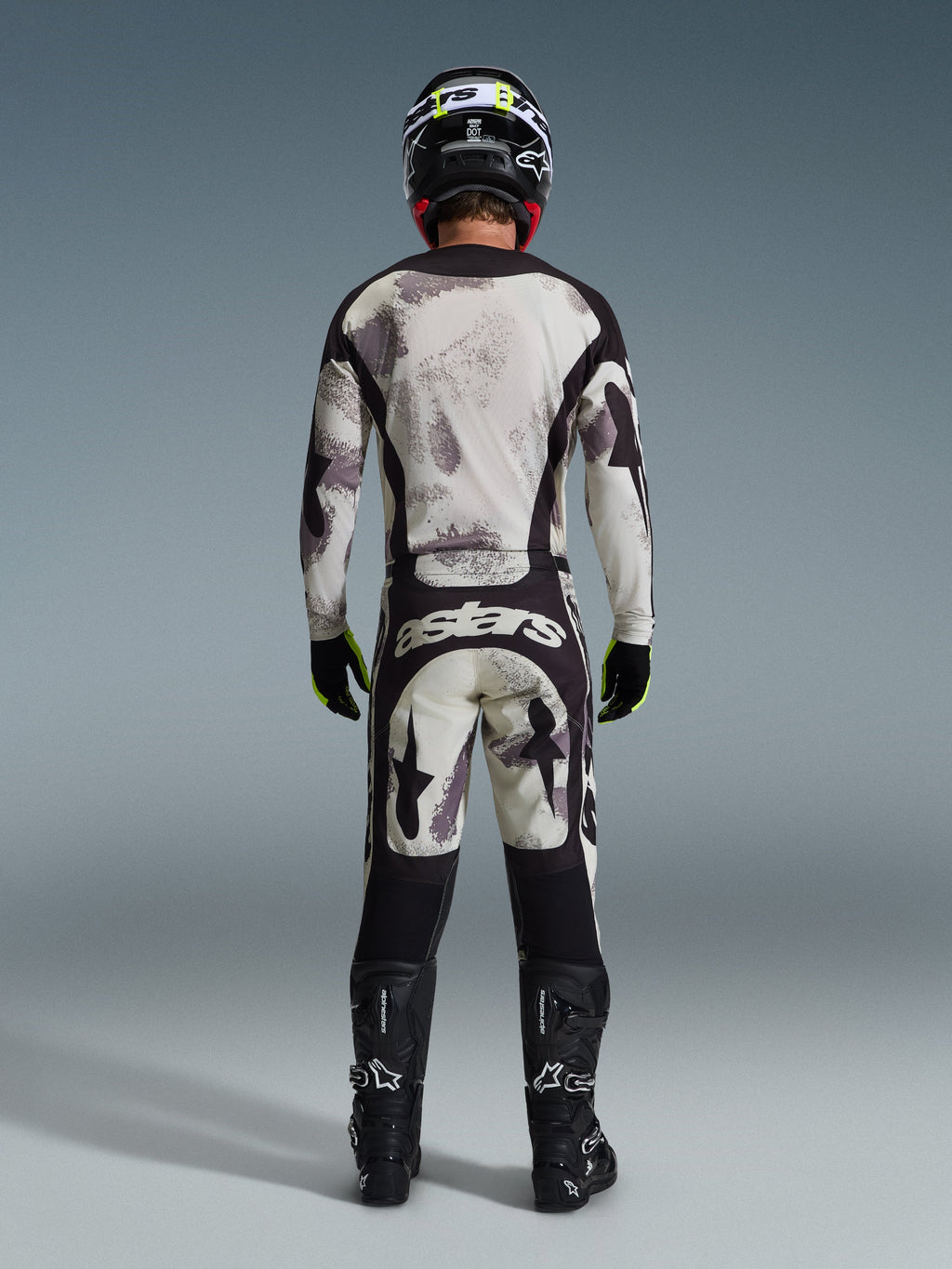Motocross rider from behind wearing the 2026 Racer Lahnd Jersey and matching pants in Sand Camo, featuring a cream and grey abstract pattern with black panels, 'astars' branding on the pants, paired with black Alpinestars boots and a black helmet for a complete off-road racing look.