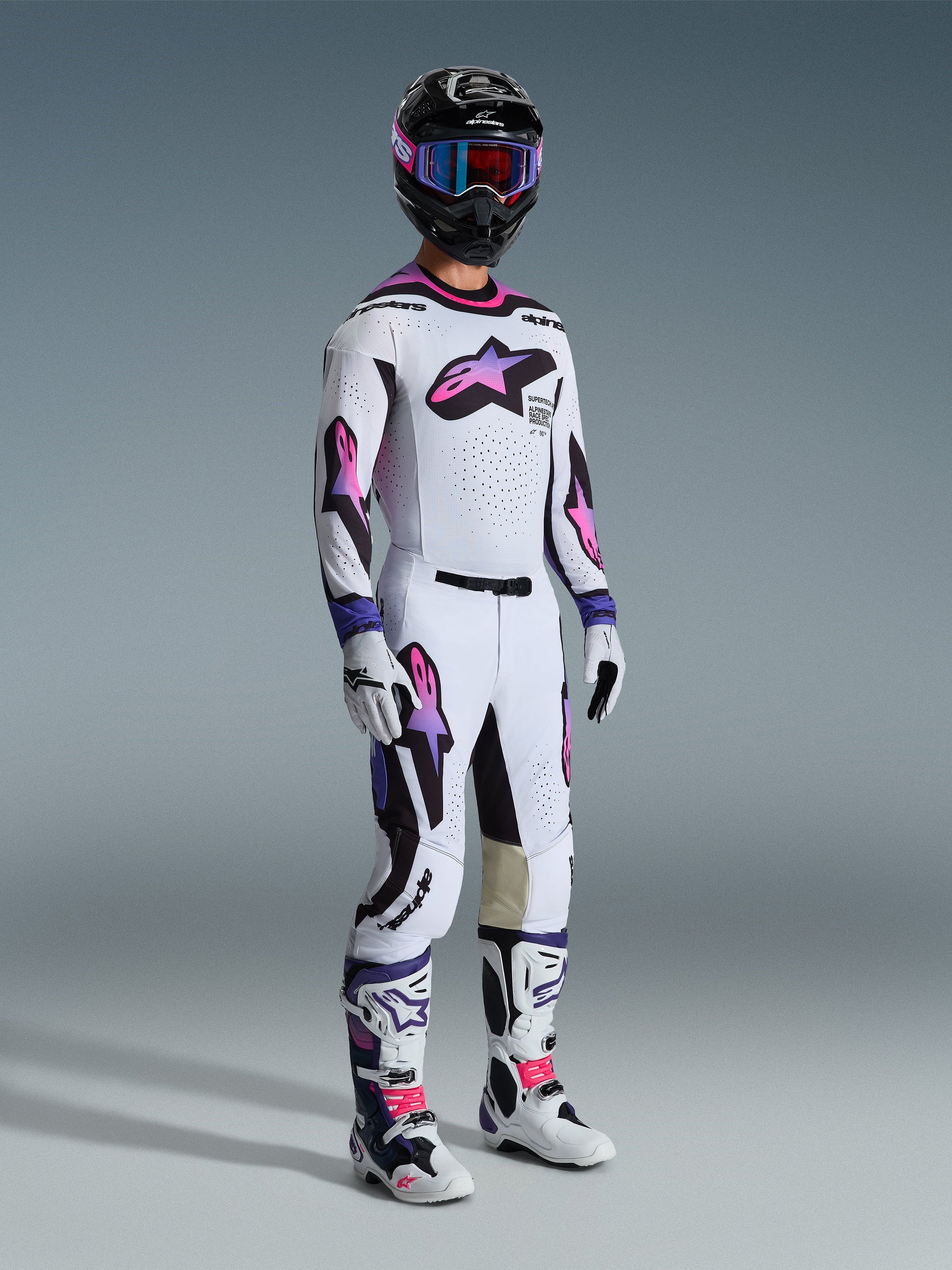 Motocross rider wearing a complete Alpinestars racing kit, featuring 2026 Supertech AFD Vista Pants in White Purple Black with matching jersey, black helmet with purple goggles, white and purple boots, and white gloves, coordinated professional off-road racing gear.