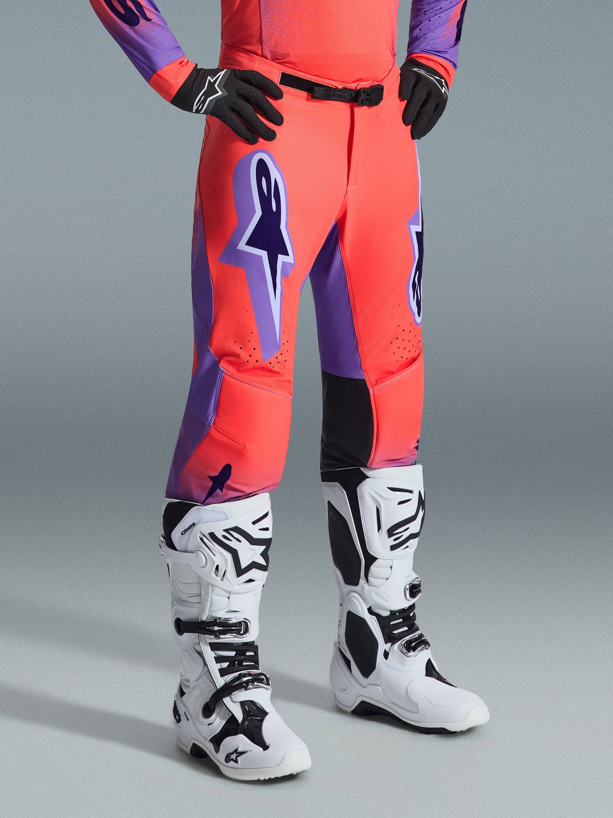 Alpinestars 2026 Supertech Scenz Pants, Motocross Pants, Hot Coral and Purple, worn with white and black motocross boots and black gloves, featuring a large purple Alpinestars logo, black leather inner knee panels, and athletic race fit design