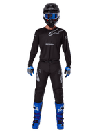 Alpinestars 2025 Racer Graphite Pants, Motocross Pants, Black and Gray, worn on a model with a matching black jersey, blue and purple boots, and blue goggles, complete off-road racing outfit featuring bold Alpinestars branding