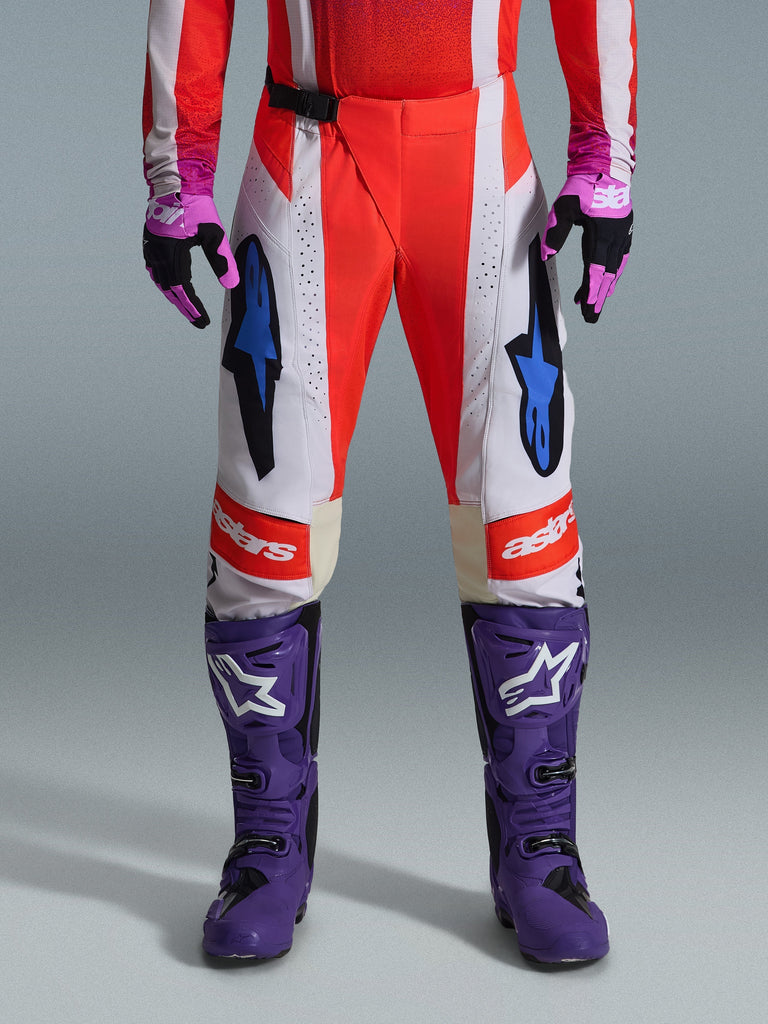 Alpinestars 2026 Techstar Knif Pants, Motocross Pants in Orange Fluo and Light Gray with Black accents, shown with purple motocross boots and gloves, featuring blue star logos and multi-panel construction for off-road performance