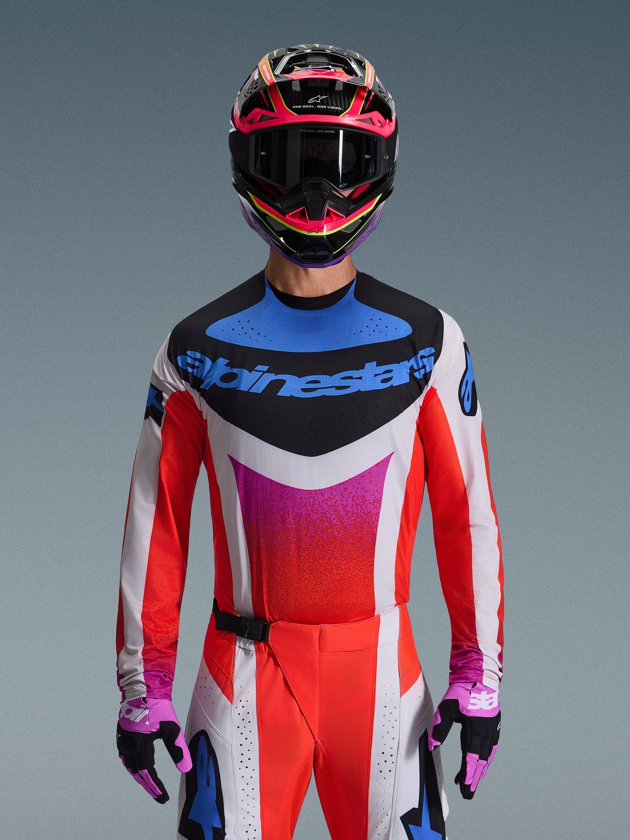 Motocross rider wearing the Alpinestars 2026 Techstar Knif Jersey in Light Gray Orange Fluo Black, featuring a bold blue logo and vibrant orange sleeves, worn with a pink and black helmet and purple gloves, athletic race fit for high-performance off-road competition