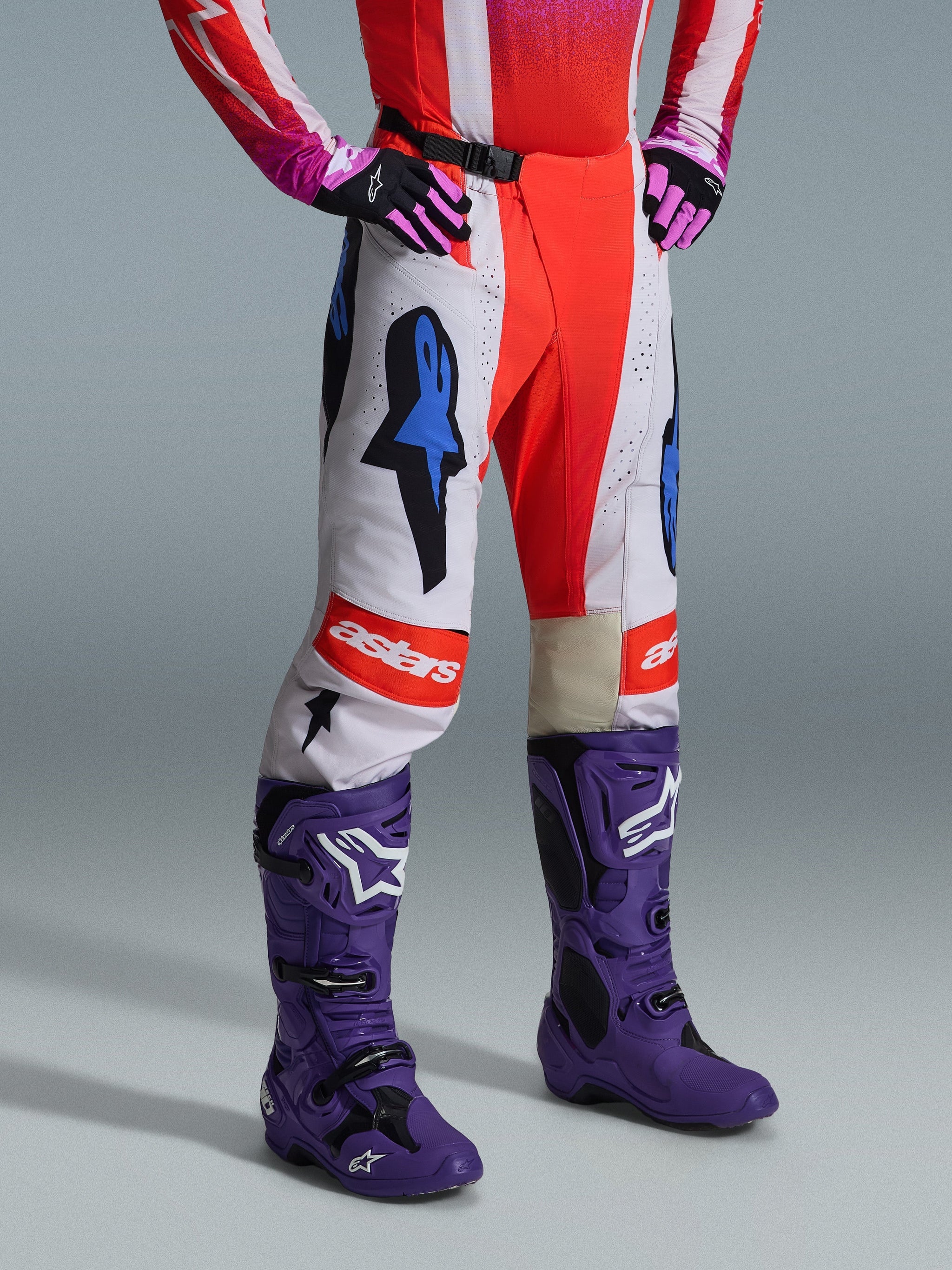 Alpinestars 2026 Techstar Knif Pants, Motocross Pants, White and Bright Red with Blue graphic accents, featuring an "astars" logo on the knee, worn with purple boots and black and pink gloves, professional off-road racing gear set.