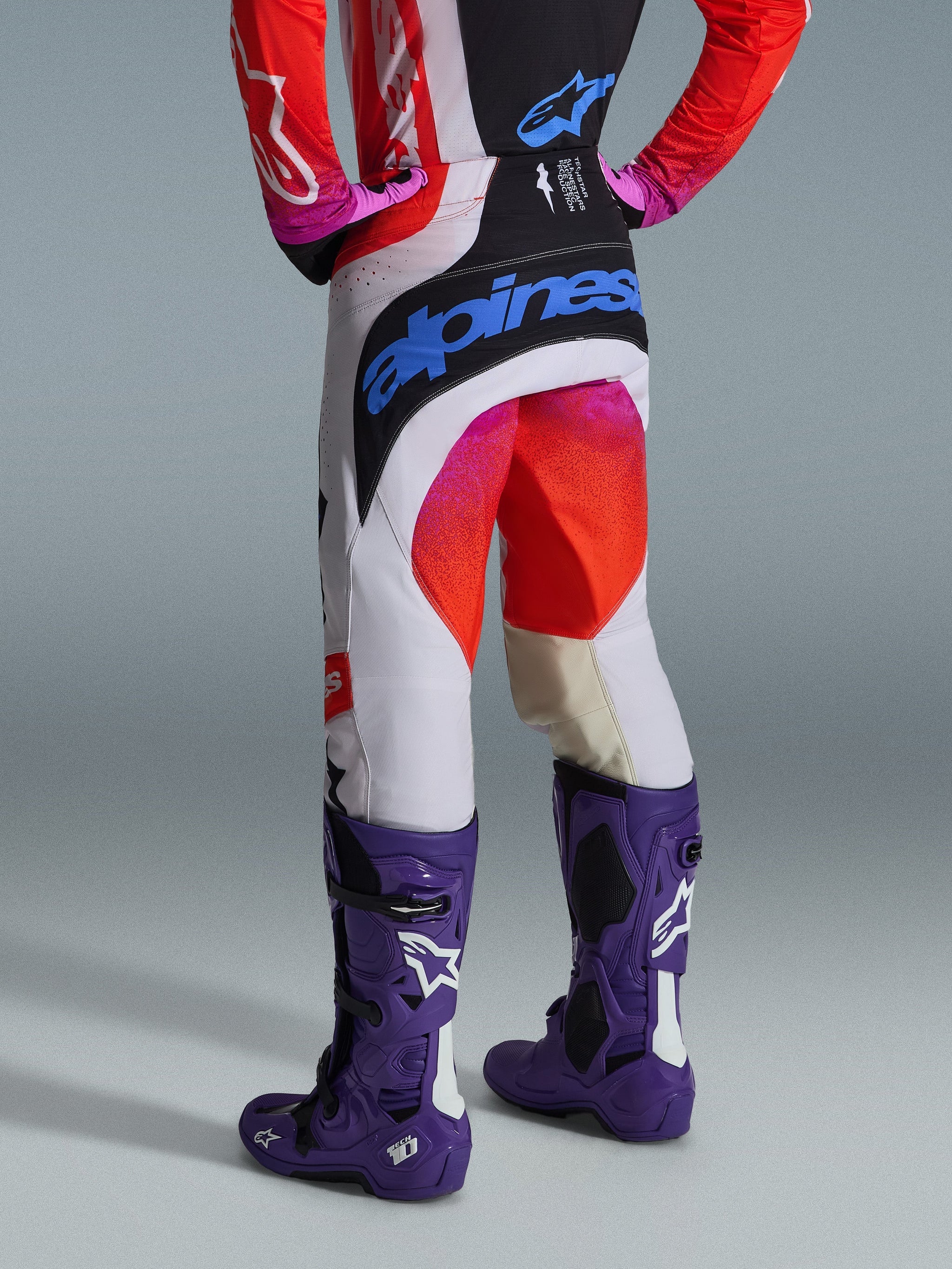 Alpinestars 2026 Techstar Knif Pants, Motocross Pants, Light Gray Orange Fluo and Black, rear view showing large blue Alpinestars branding on the seat, worn with purple Tech 10 boots, professional off-road racing gear.