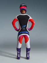 Motocross rider wearing complete 2026 Techstar Knif racing gear from the back, light grey jersey with black back panel and orange sleeves, matching white and orange pants featuring blue Alpinestars branding, purple boots and a black and pink helmet, coordinated professional outfit for off-road racing