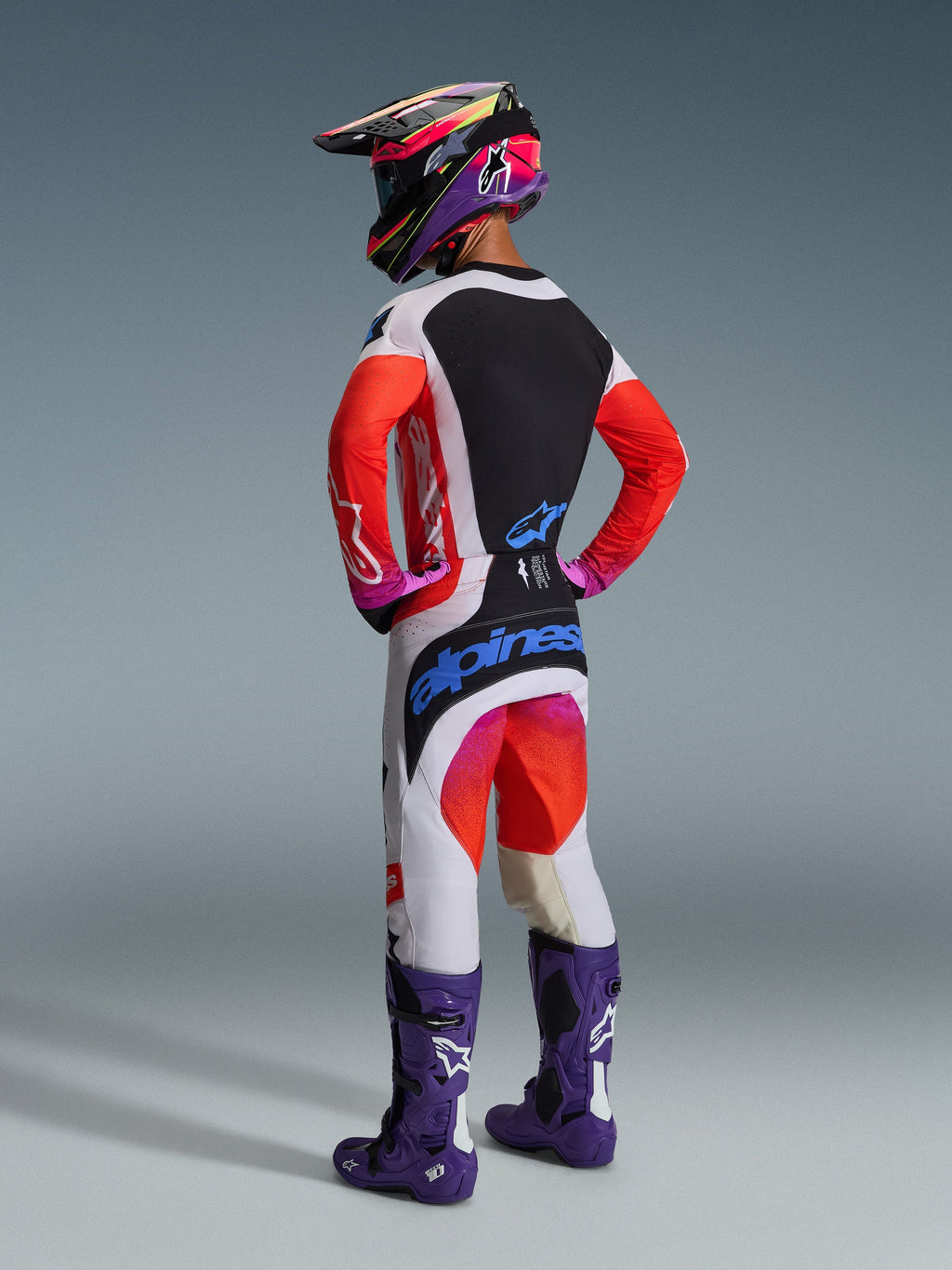 Motocross rider viewed from behind wearing complete racing gear, Alpinestars 2026 Techstar Knif Pants in Light Gray and Orange Fluo with blue branding, matching jersey with orange sleeves, purple Tech 10 boots, and a colorful racing helmet, coordinated professional off-road outfit