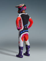 Rear view of a motocross rider wearing the Alpinestars 2026 Techstar Knif Jersey, light gray and black with fluorescent orange sleeves, paired with matching racing pants, purple boots, and a multi-colored helmet, professional off-road gear set.
