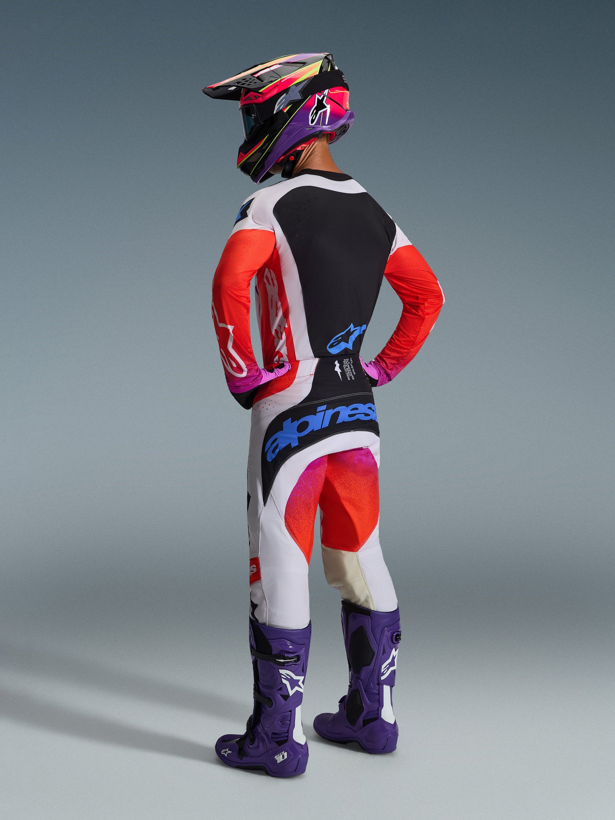 Rear view of a motocross rider wearing the Alpinestars 2026 Techstar Knif Jersey, light gray and black with fluorescent orange sleeves, paired with matching racing pants, purple boots, and a multi-colored helmet, professional off-road gear set.