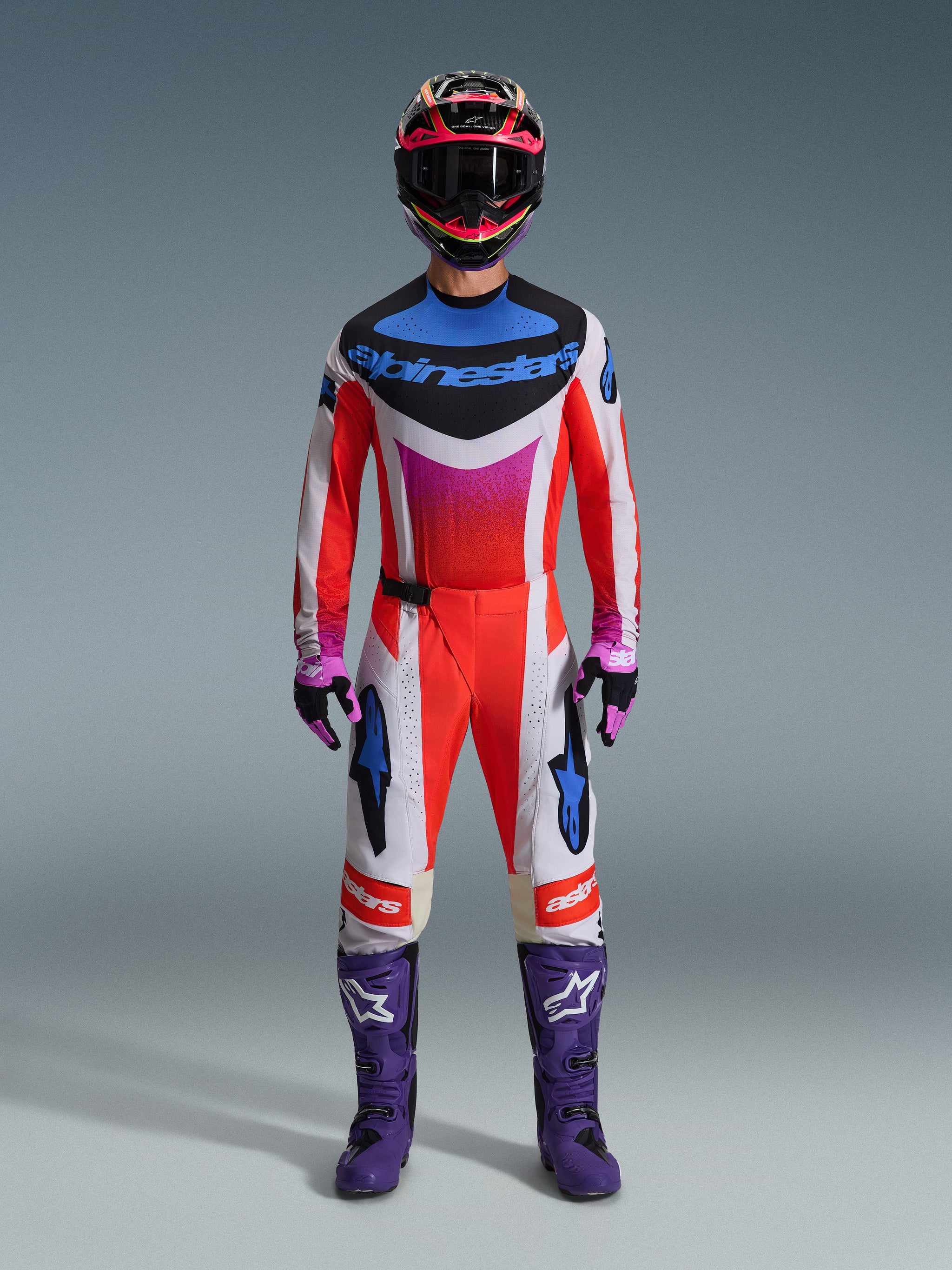 Motocross rider wearing complete racing gear, Alpinestars 2026 Techstar Knif Jersey and matching pants in white and orange with blue and purple accents, purple racing boots with white logo, and a multi-colored helmet, coordinated outfit for off-road racing competition