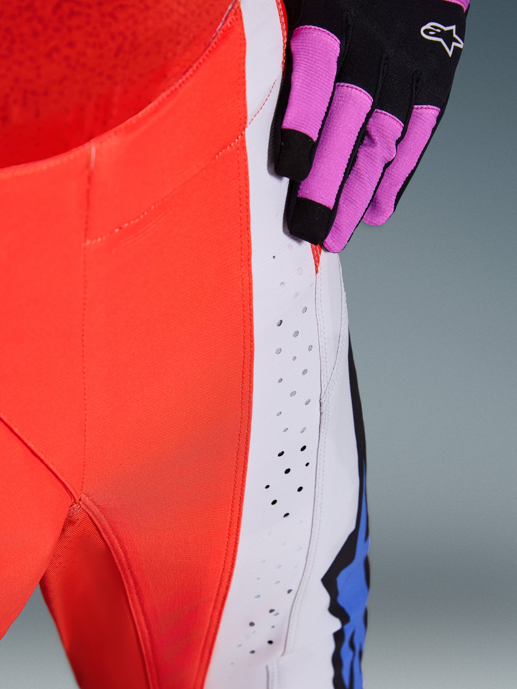 Alpinestars 2026 Techstar Knif Pants, Motocross Pants, Orange Fluo and Light Gray with blue accents, close-up showing laser-cut ventilation perforations on the thigh, worn with a black and purple motocross glove