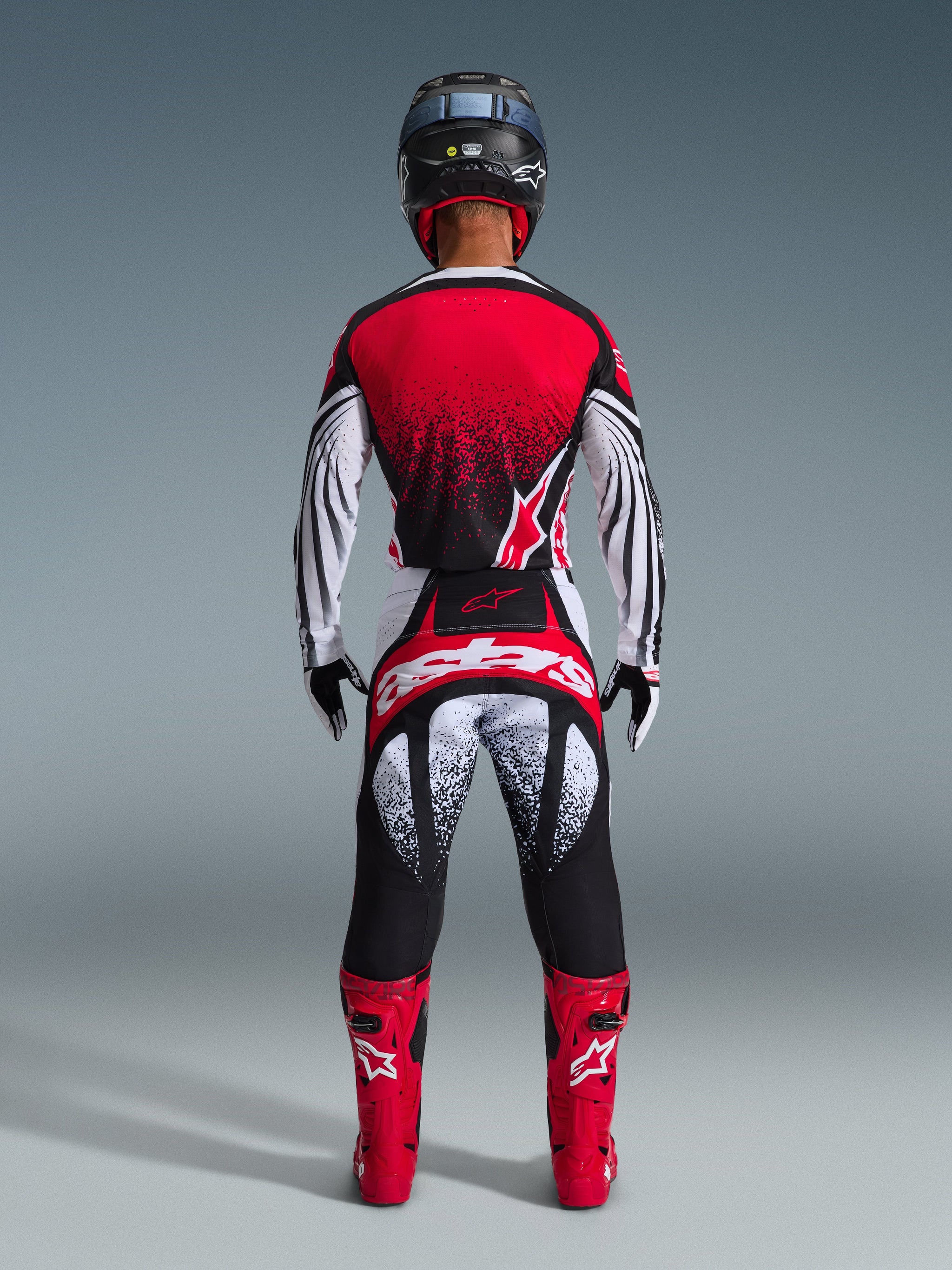 Rear view of a motocross rider wearing Alpinestars 2026 Techstar Nomur Pants in Black White Red, featuring bold branding on the seat and speckled graphics, paired with a matching red jersey and bright red boots for a complete off-road racing kit