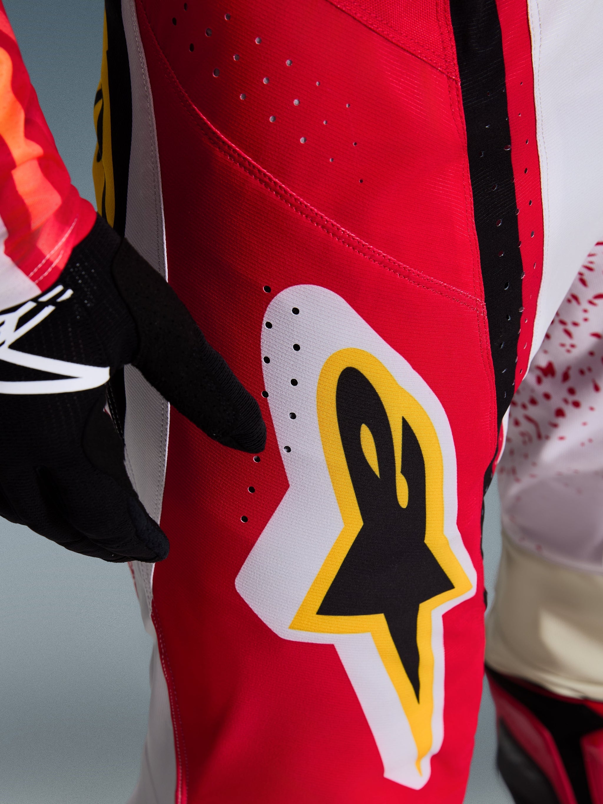 Alpinestars 2026 Techstar Nomur Pants, Motocross Pants, Red with white and yellow accents, close-up of thigh area featuring laser-cut ventilation holes and a large star logo, paired with black gloves and a colorful jersey for off-road performance.