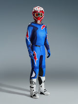 Motocross rider wearing complete racing gear, Blue and Red jersey with grid pattern and Alpinestars logo, 2026 Fluid Grid Pants in Blue Red with black inner knee panels, red and white helmet with goggles, white and black boots, blue gloves, coordinated outfit for off-road racing
