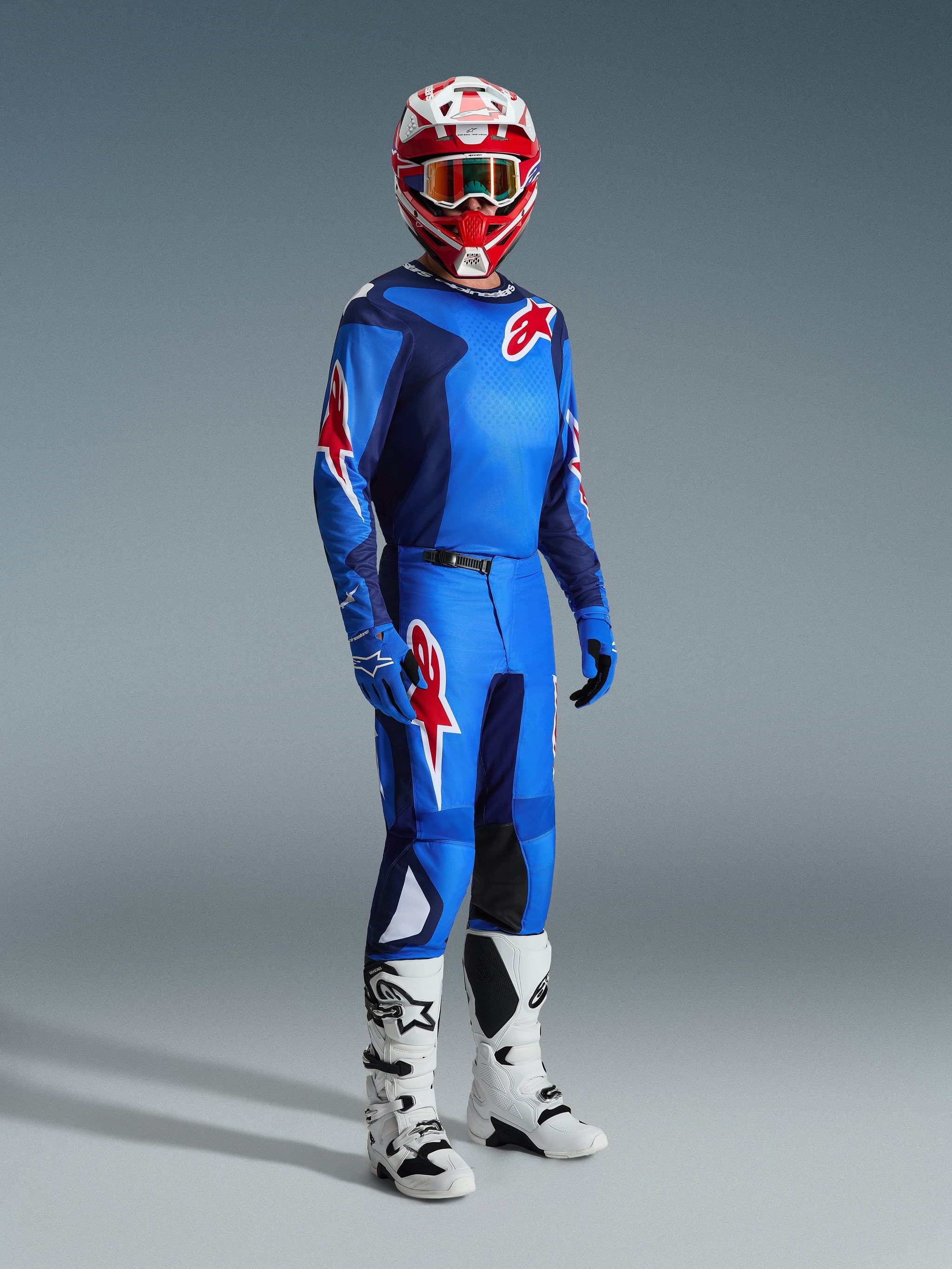 Motocross rider wearing complete racing gear, Blue and Red jersey with grid pattern and Alpinestars logo, 2026 Fluid Grid Pants in Blue Red with black inner knee panels, red and white helmet with goggles, white and black boots, blue gloves, coordinated outfit for off-road racing
