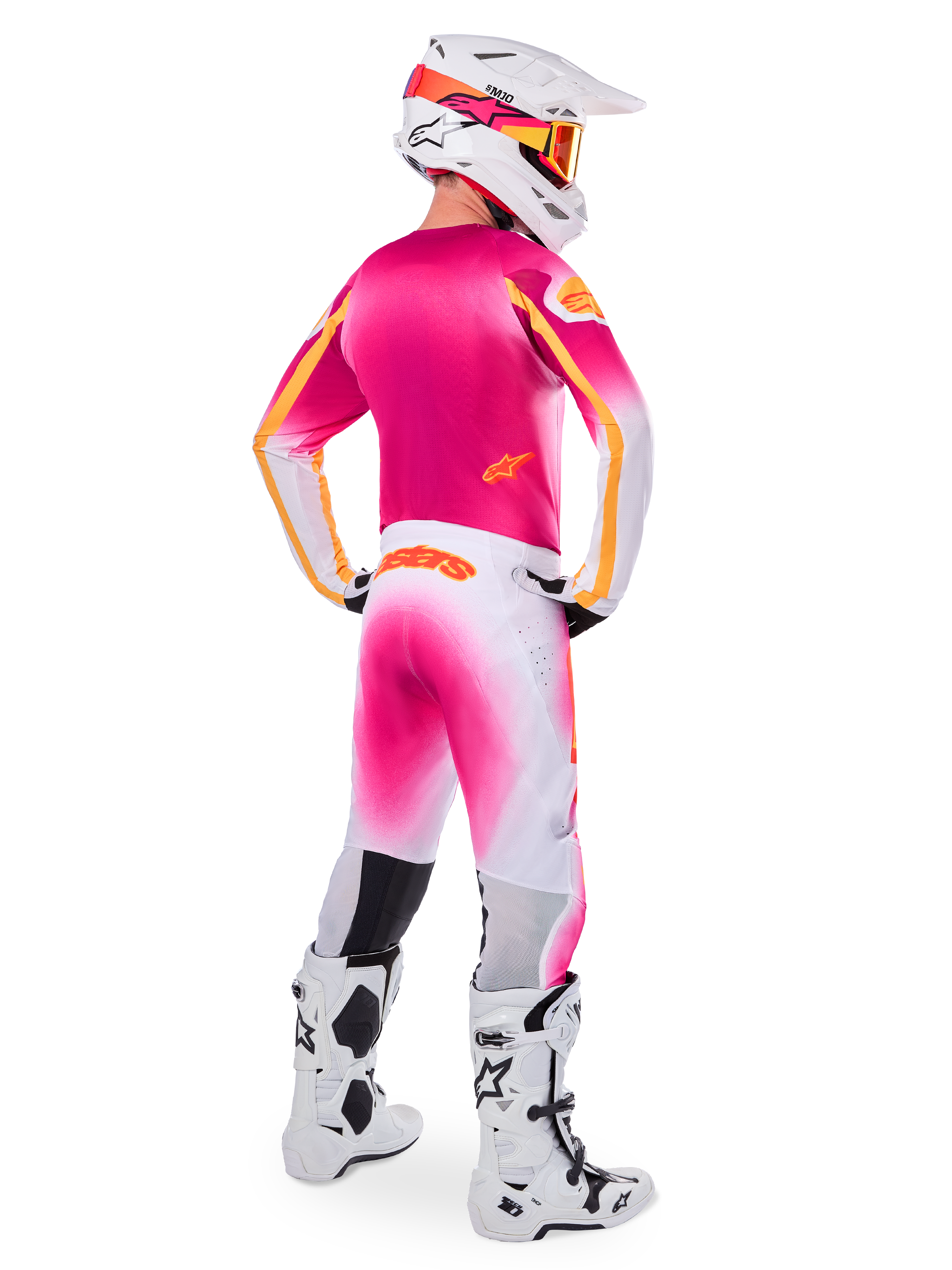 Motocross rider seen from the back wearing the Limited Edition Supertech AFD MIG Gear Set, pink jersey with orange accents, white pants with pink gradient and orange Alpinestars branding, white helmet with pink graphics and orange goggles, white and black boots, professional off-road racing kit