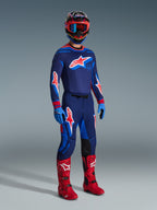 Motocross rider wearing 2026 Supertech Vista Pants in Dark Navy Hot Coral Cobalt, matching long-sleeve racing jersey, red motocross boots, and coordinated helmet with red goggles, full-body studio shot of high-performance off-road gear
