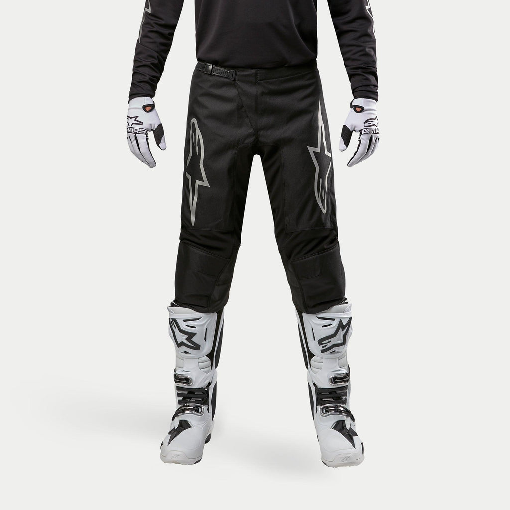Alpinestars 2024 Fluid Graphite Pants, Motocross Pants, Black and Silver, featuring silver Alpinestars logos on the thighs, worn with white and black motocross boots and gloves, sporty design for off-road racing