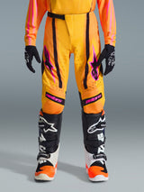 Alpinestars 2026 Youth Supertech Nomur Pants, Motocross Pants, Orange Yellow and Pink, worn with matching jersey and boots, featuring black stripes, pink star logos, and perforated zones, designed for competitive youth motocross racing