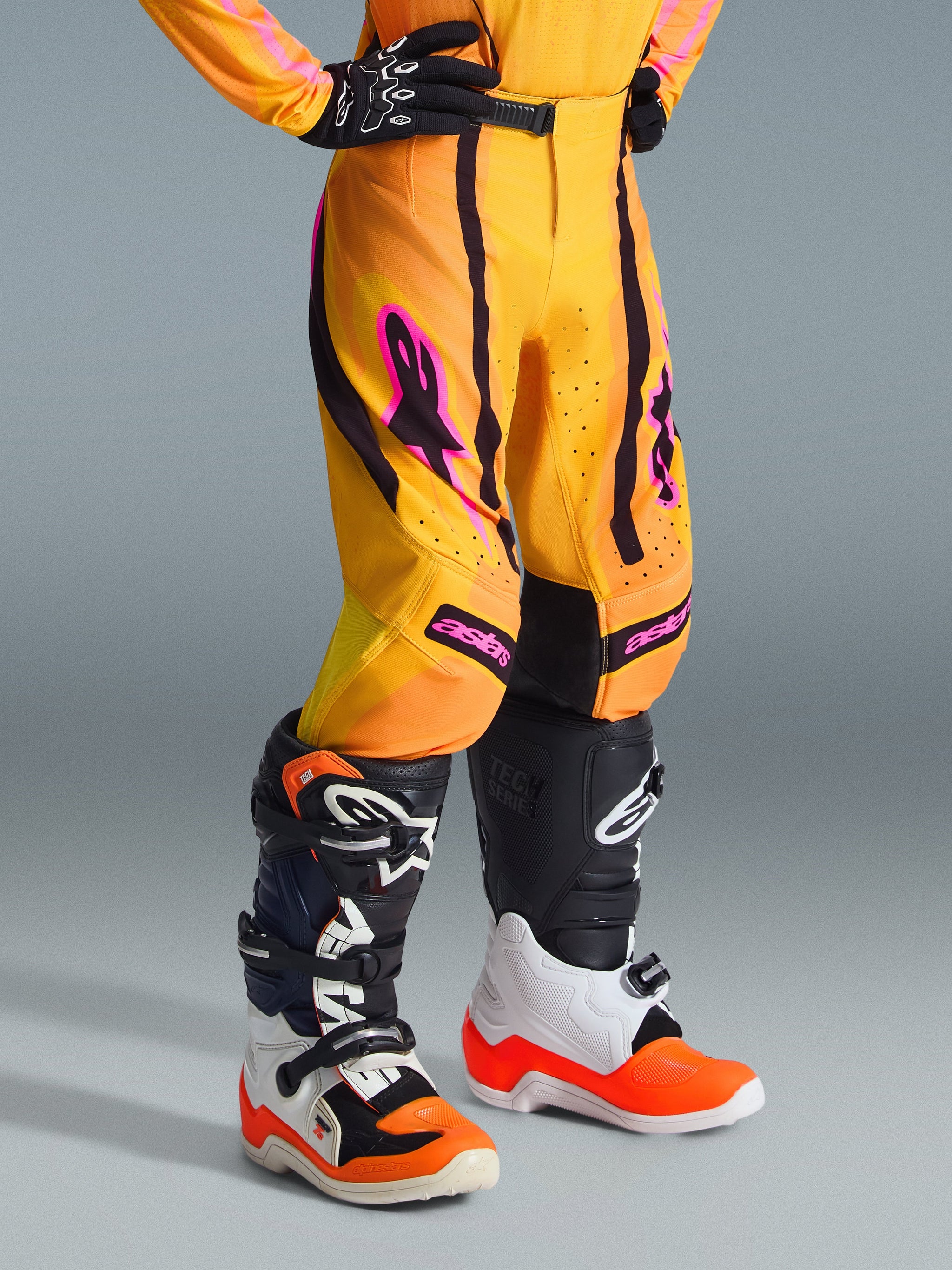 Alpinestars 2026 Youth Supertech Nomur Pants, Motocross Pants, Orange Yellow Pink, worn with black gloves and black and white motocross boots, featuring vibrant pink logo accents and black panels, athletic race fit for off-road riding.