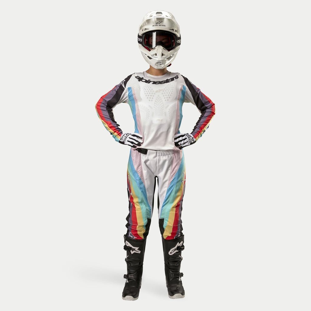 Female motocross rider wearing Alpinestars Women Stella 2024 Techstar Pants, Black Multicolor featuring white base with red, yellow, and blue stripes, matching long-sleeve jersey, white helmet, and black boots, full racing gear for off-road competition