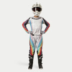 Female motocross rider wearing Alpinestars Women Stella 2024 Techstar Pants, Black Multicolor featuring white base with red, yellow, and blue stripes, matching long-sleeve jersey, white helmet, and black boots, full racing gear for off-road competition