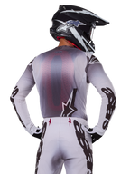Alpinestars 2025 Supertech Maker Gear Set, motocross rider shown from the back, white and gray jersey with large black branding on sleeves and star logo, matching white pants with black graphics, black and white helmet, professional off-road racing outfit