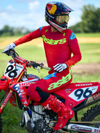 Motocross rider on a red Honda motorcycle wearing the Alpinestars 2025 Supertech Maker Gear Set in Red, featuring a red jersey with neon green 'astars' branding and blue shoulder accents, matching red pants, solid red boots, white gloves, and a Red Bull helmet, professional racing kit for off-road performance