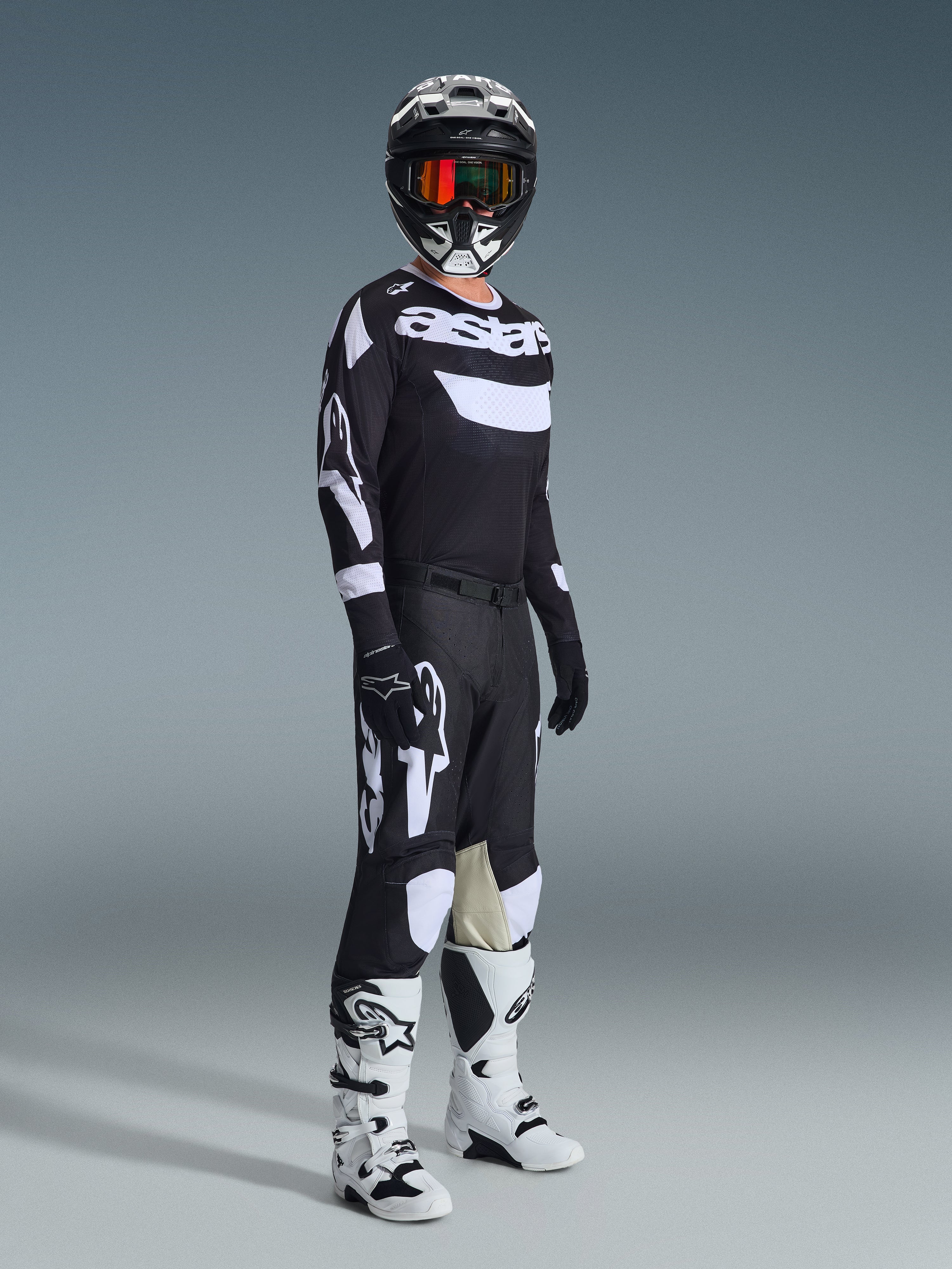 Motocross rider wearing complete racing gear, 2026 Racer Air Riway Jersey in Black and White, matching black pants with large white Alpinestars logos, black helmet with white accents and orange goggles, white and black boots, coordinated outfit for professional off-road racing