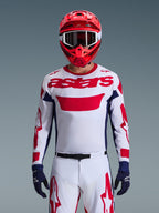Motocross rider wearing the Alpinestars 2026 Racer Riway Jersey in White Blue Red, featuring a large red 'astars' logo on the chest, navy blue side panels, matching white and red motocross pants, a red helmet with white goggles, and navy blue gloves, complete off-road racing gear set.