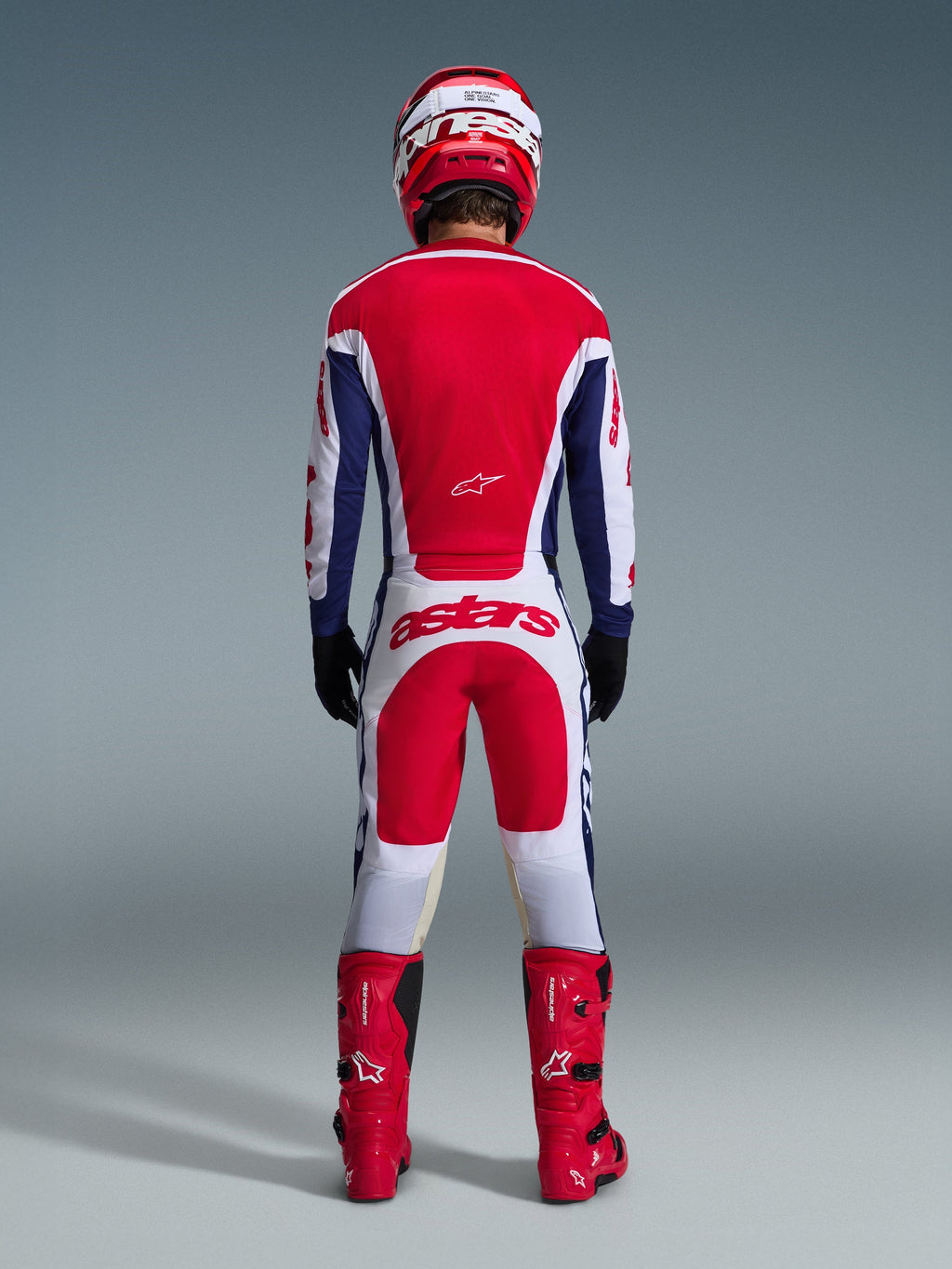 Motocross rider viewed from behind wearing the 2026 Racer Riway Gear Set, Red and blue jersey with white accents, white pants with red panels and 'astars' logo on the seat, coordinated with a red helmet, red boots, and black gloves, high-performance racing gear for off-road